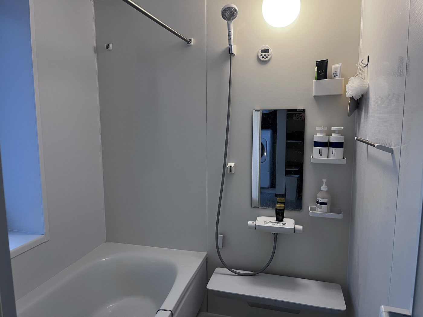 A Japanese unit bathroom: a waterproof room with a tub, a wall-mounted shower, a mirror and small wall-mounted racks holding bottles
