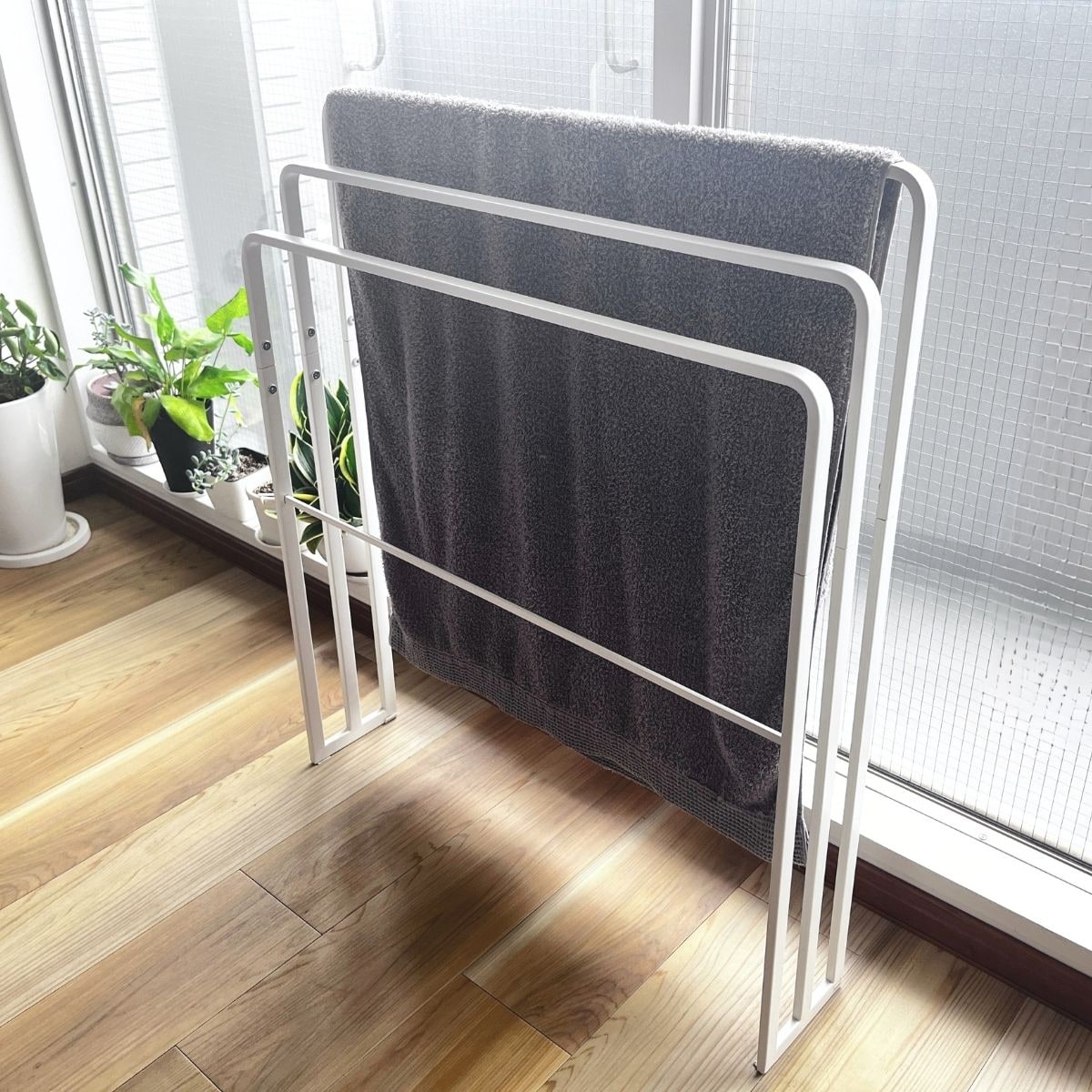 A white freestanding towel stand with three bars and a bath mat bar, holding a gray bath towel by a window