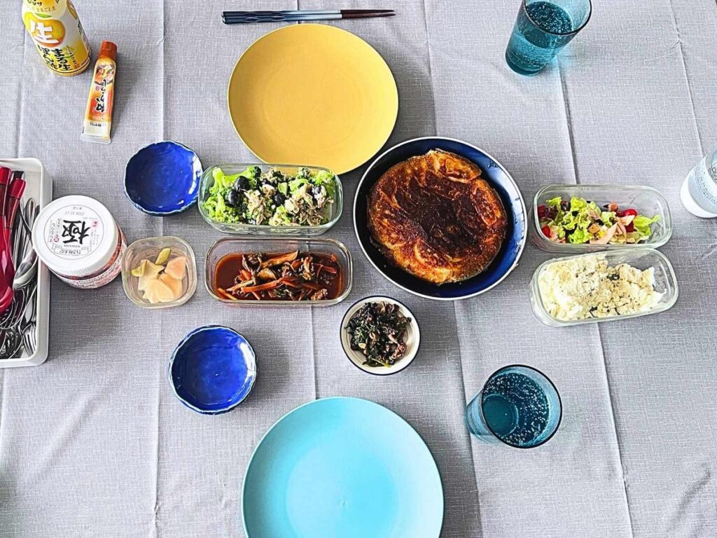 A Japanese home lunch on the table, seen from above: gyoza on a blue plate, a broccoli salad and leftovers in glass containers, and a jar of kimchi