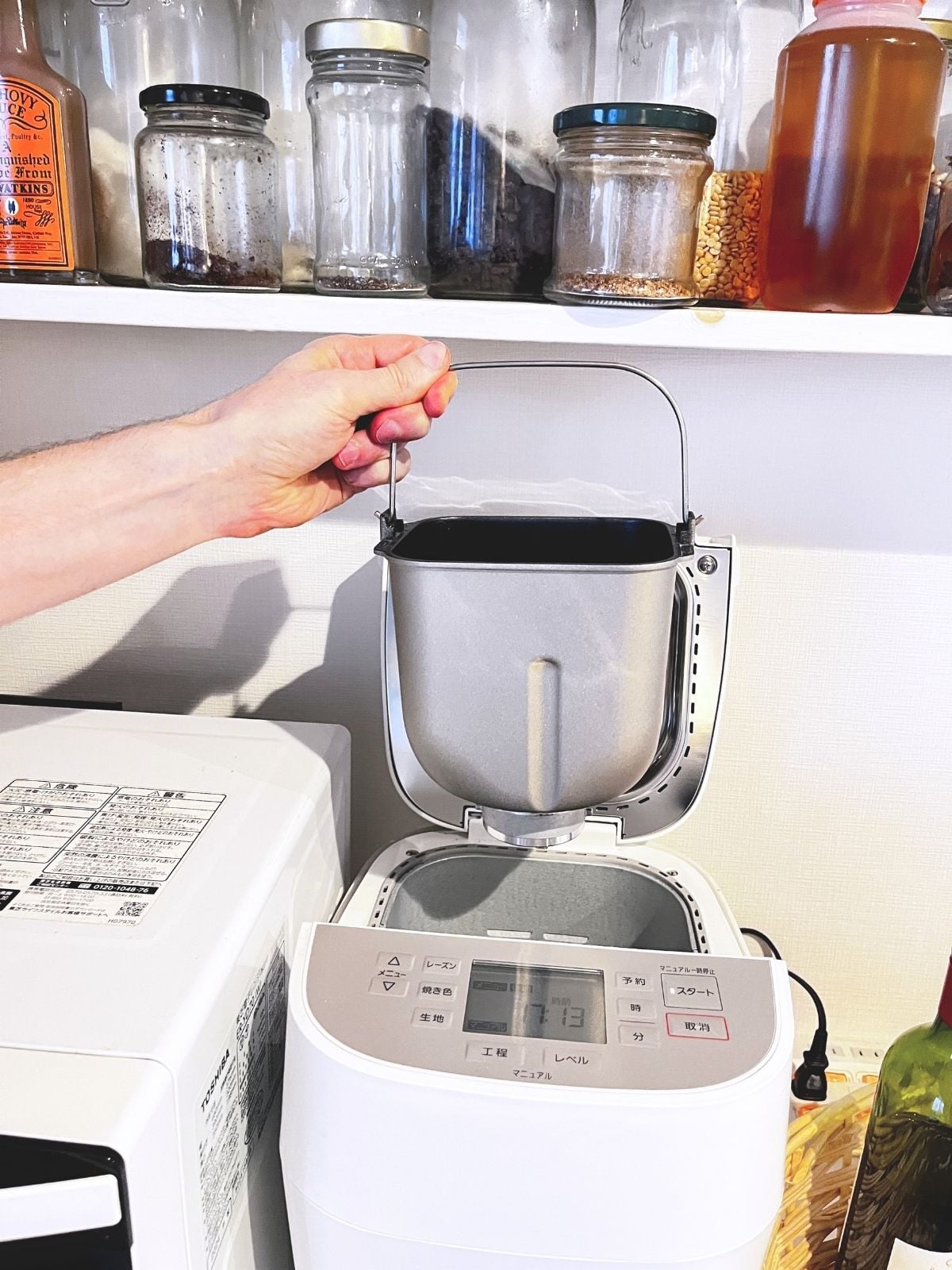 A bread maker with its lid fully open under the lower shelf, the bread pan being lifted straight out