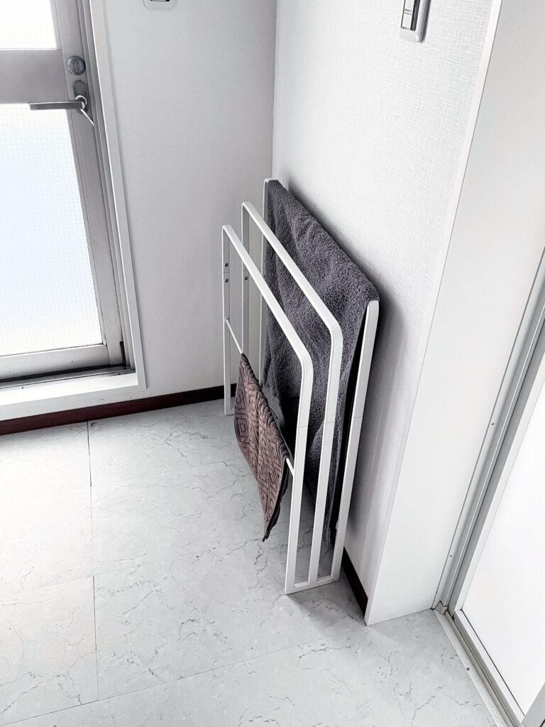 A white freestanding towel stand holding a bath towel and a bath mat, set against the wall beside a door in a Japanese washroom
