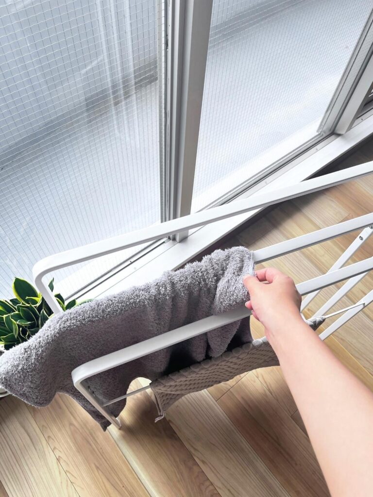 A hand sliding a gray towel onto a freestanding towel rack from the side, with a bath mat on the lower bar
