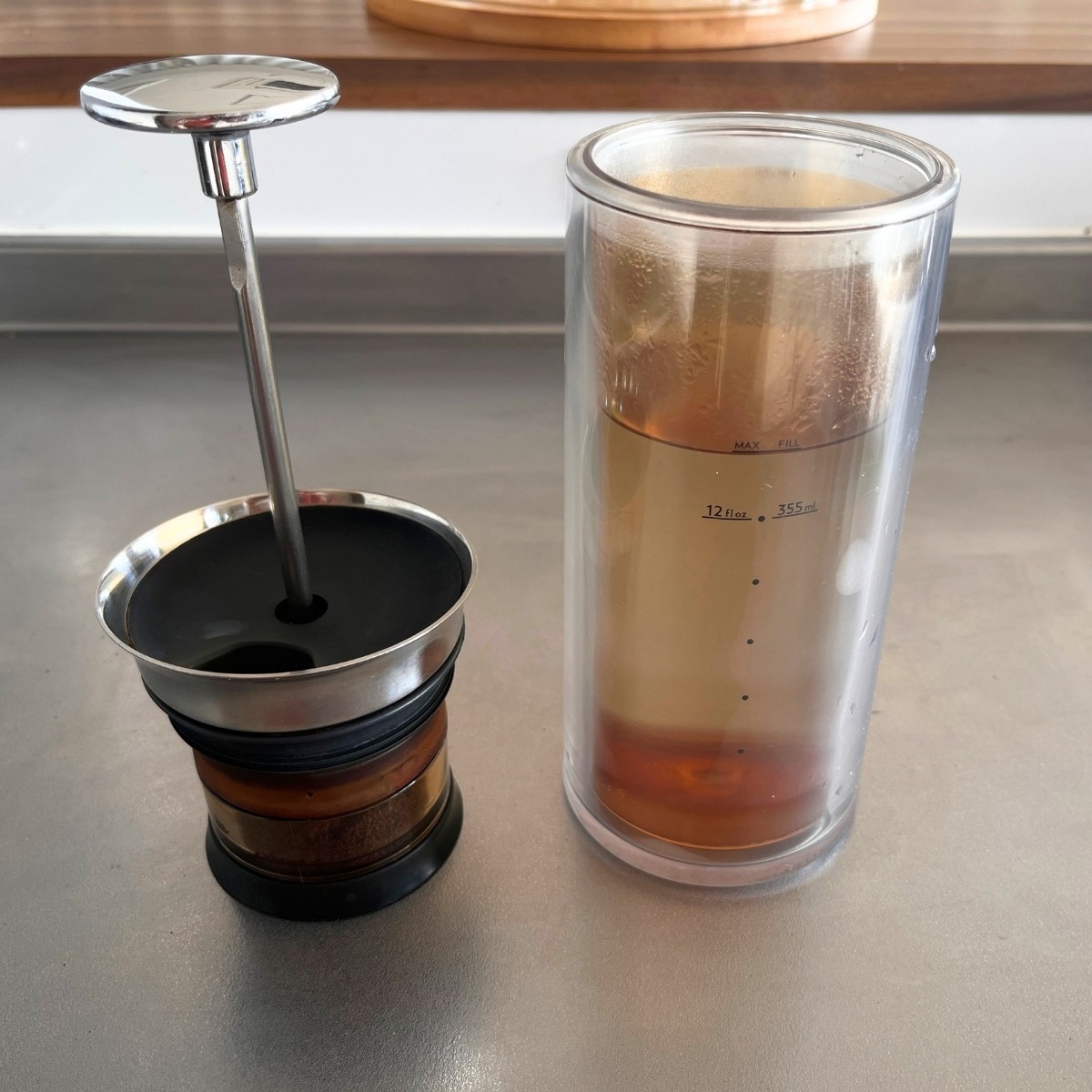 American Press coffee maker plunger with the coffee-filled pod, next to the carafe of hot water on a stainless steel counter