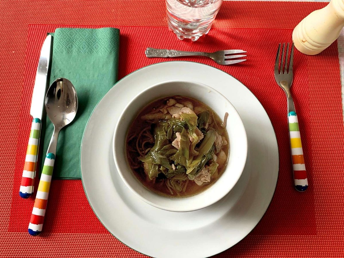 A bowl of soba with chicken and leeks on a red placemat
