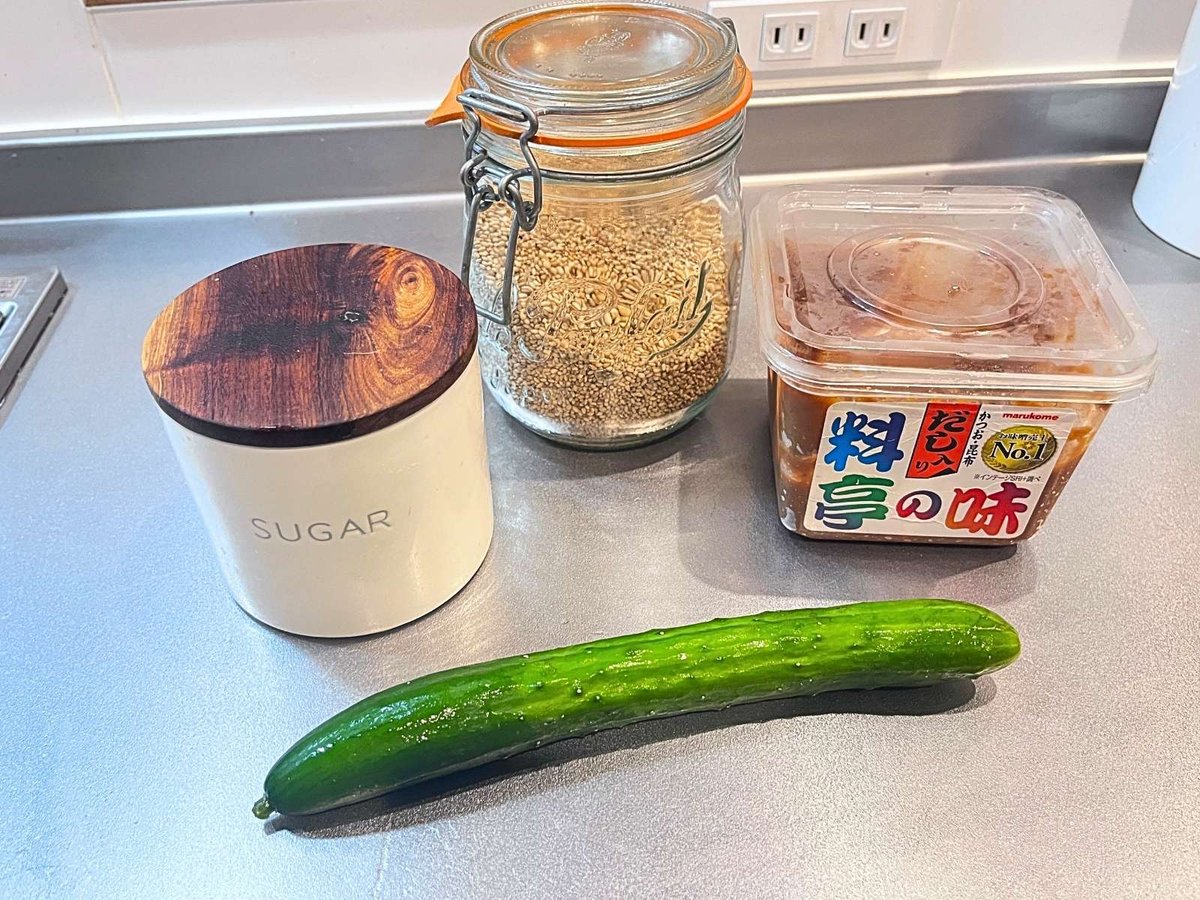 A cucumber, a tub of miso, a jar of sesame seeds and a sugar canister on a kitchen counter