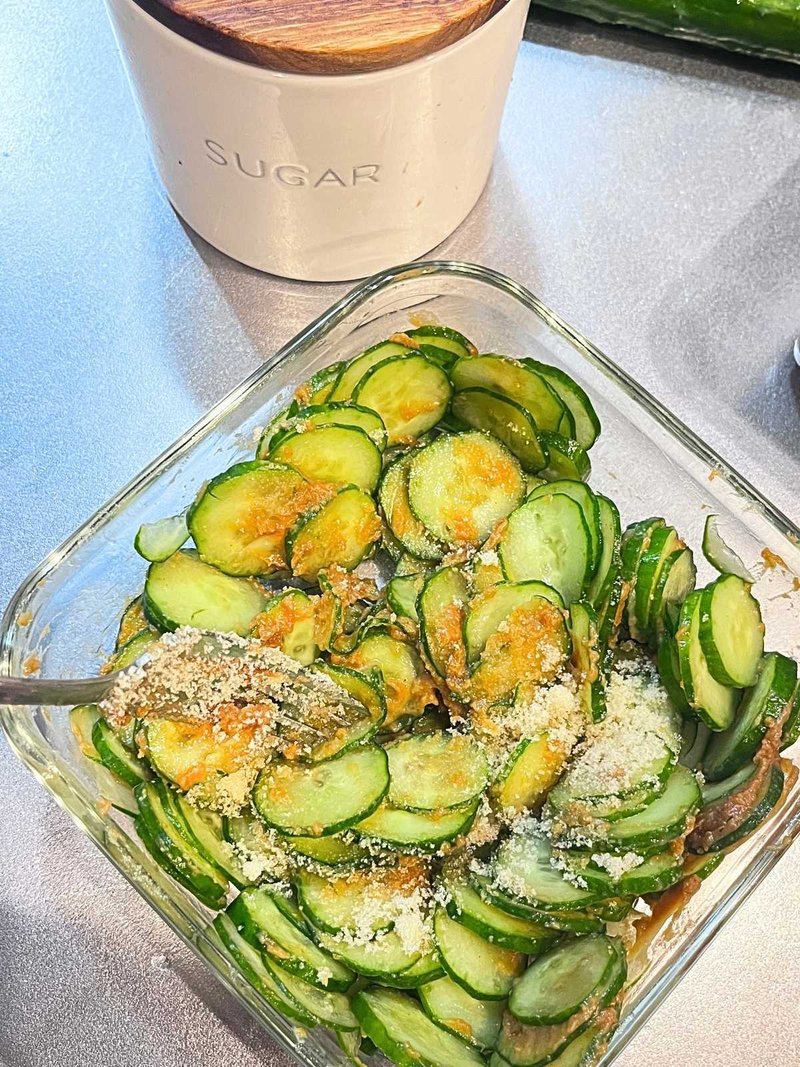 Sugar being mixed into sliced cucumber and miso with a fork in a square glass container