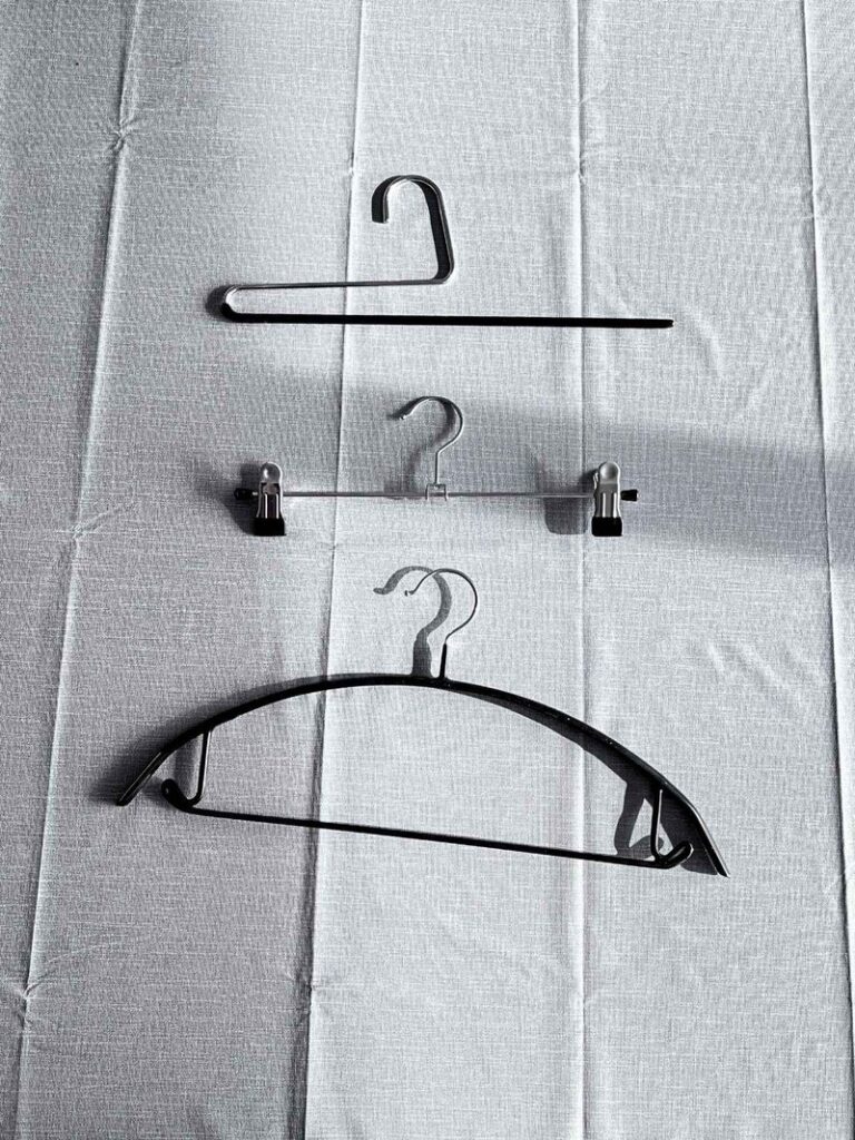 Three hangers laid out on a table: a single bar hanger, a pants hanger with two sliding clips, and a black non-slip hanger with shoulder notches
