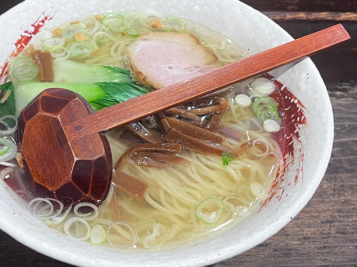 The carved back of a wooden spoon resting on a bowl of shio ramen