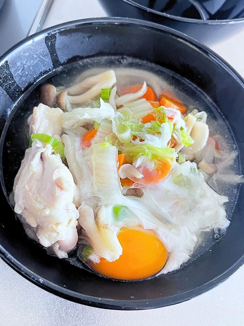 A black bowl of broth with chicken drumettes, napa cabbage, carrot, mushrooms, green onion and a raw egg yolk on top