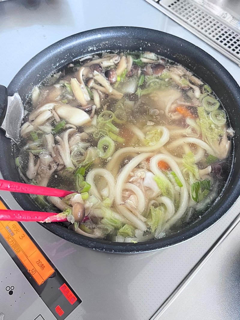 Udon, mushrooms and chicken simmering in broth with vegetables in a black pan, with red cooking chopsticks